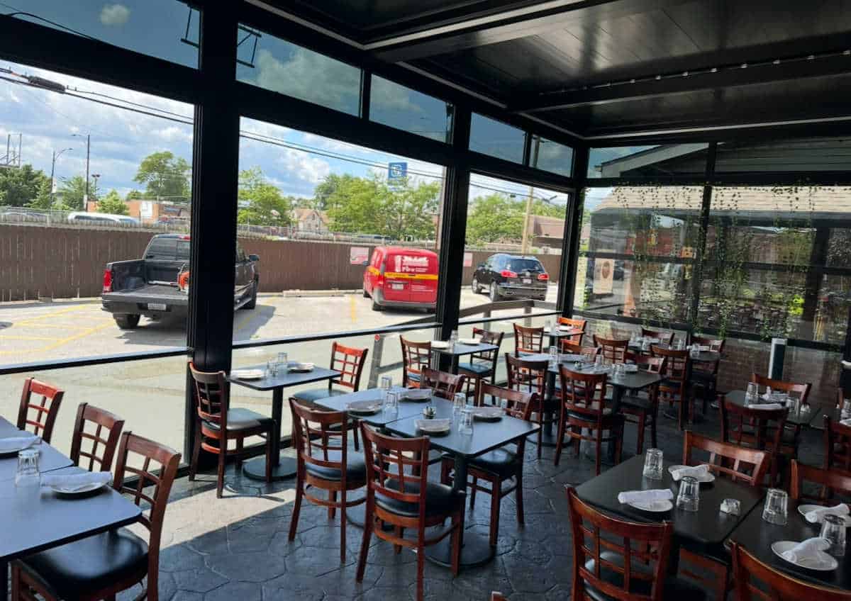 Interior view of Ciao Ragazzi’s glass-enclosed patio seating area in Chicago, showing neatly arranged tables with place settings and a view of the parking lot through large floor-to-ceiling windows.