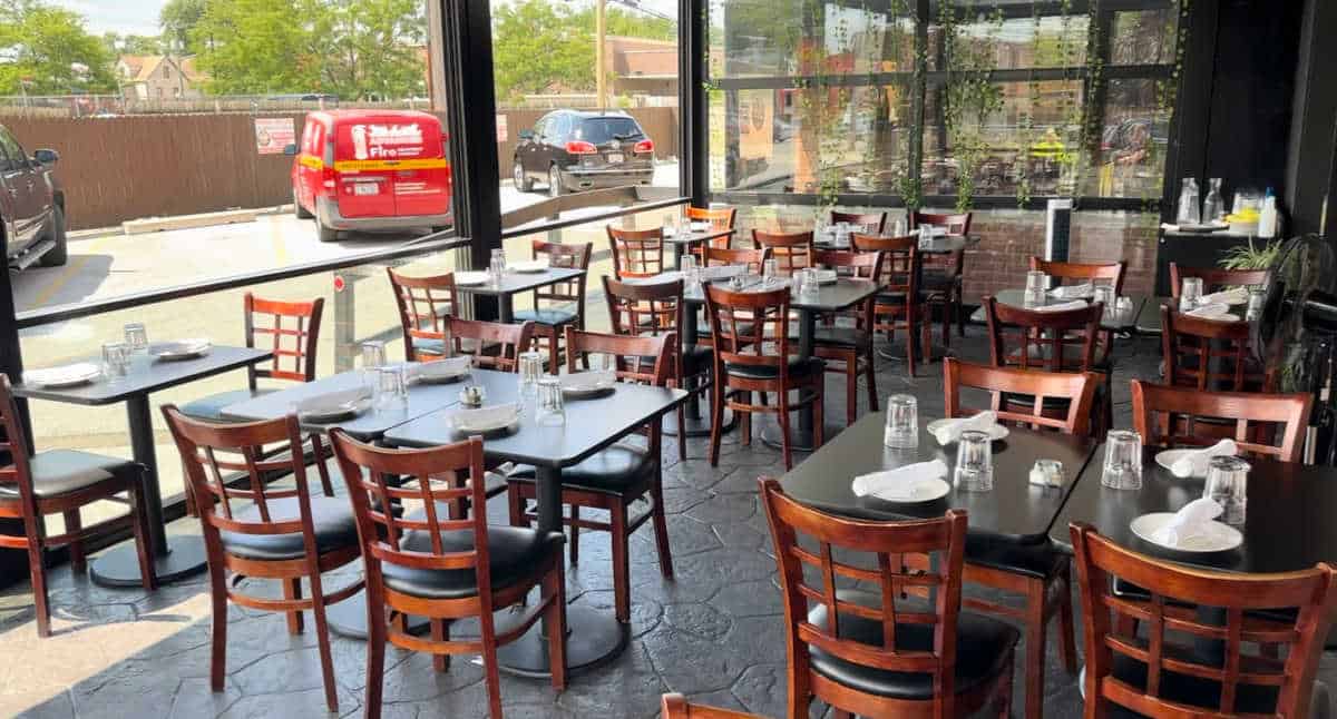 Glass-enclosed patio dining area at Ciao Ragazzi Restaurant in Chicago, featuring neatly arranged tables with wooden chairs, place settings, and natural light coming through large windows overlooking the parking lot.