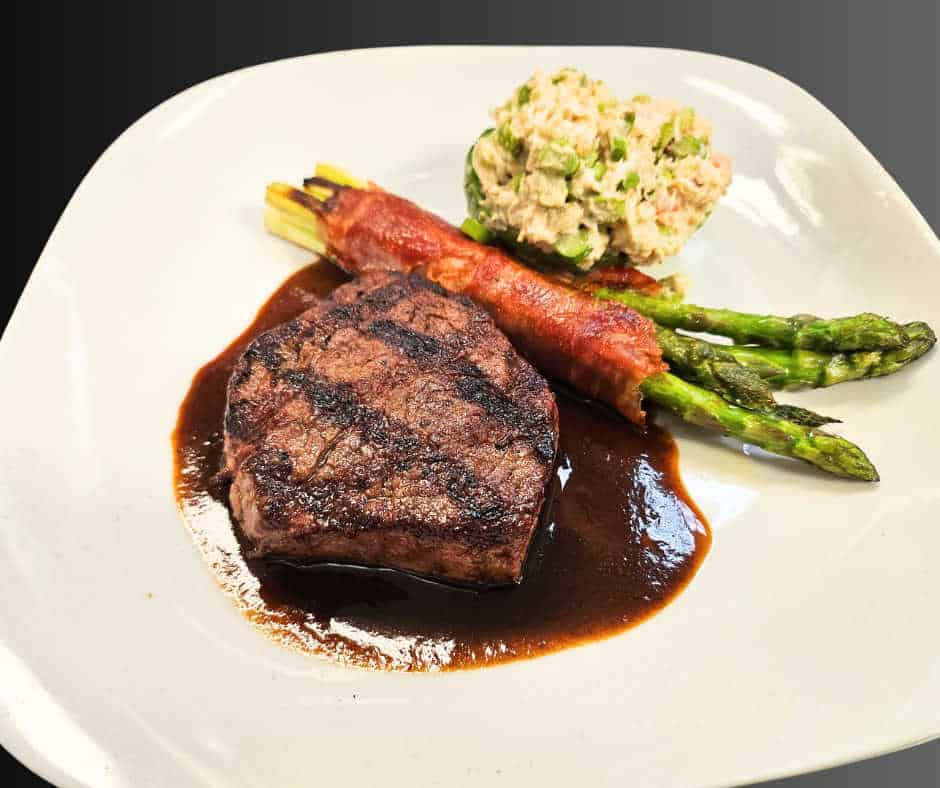 A grilled filet mignon served with a rich brown sauce, accompanied by prosciutto-wrapped asparagus and a side of creamy vegetable salad, elegantly plated on a square white dish.