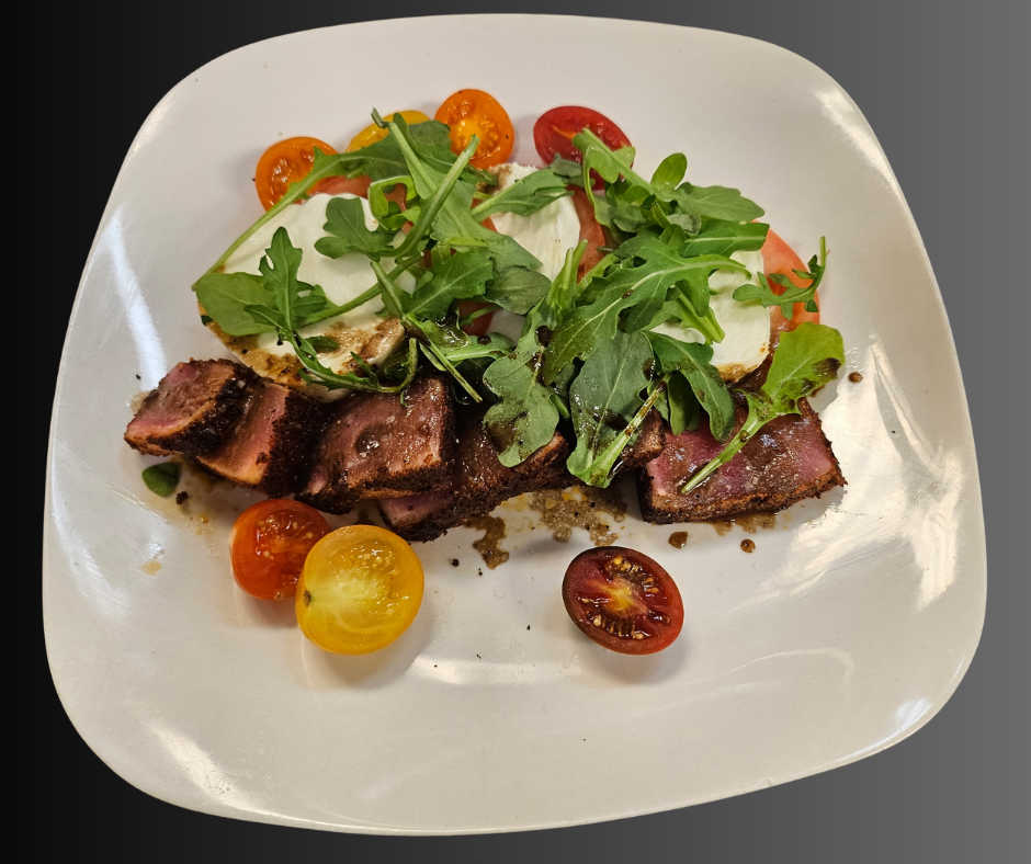 A plated dish featuring sliced seared steak topped with fresh arugula, served alongside mozzarella, heirloom cherry tomatoes, and a drizzle of balsamic glaze on a square white plate.
